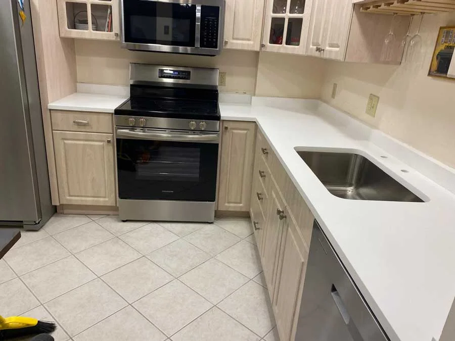 Bright L-shaped kitchen with light wood cabinets, white solid-surface countertops, stainless steel appliances, an undermount sink, and beige tiled flooring.